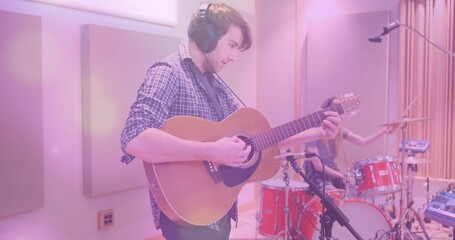 Playing guitarist in checked shirt, headphones, tracking acoustic guitar in studio, red drum kit