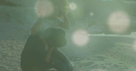 Kneeling couple adjusting hair and holding black hat on sandy shoreline, striped shirt, lens flare