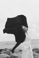 Wind sweeps a cape over a white dress as a woman stands on a rocky shore, sea and sky behind,...