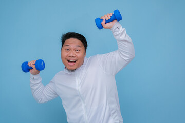 Close up of young Asian man in the gym lifting dumbbell wearing white t-shirt with happy laughing expression on turquoise blue background