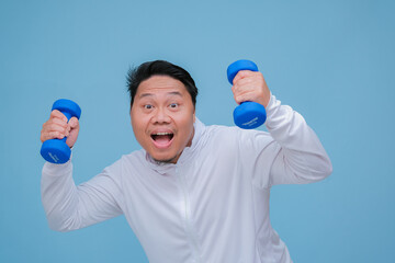 Close up of young Asian man in the gym lifting dumbbell wearing white t-shirt with happy laughing expression on turquoise blue background