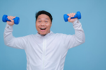 Close up of young Asian man in the gym lifting dumbbell wearing white t-shirt with happy laughing expression on turquoise blue background