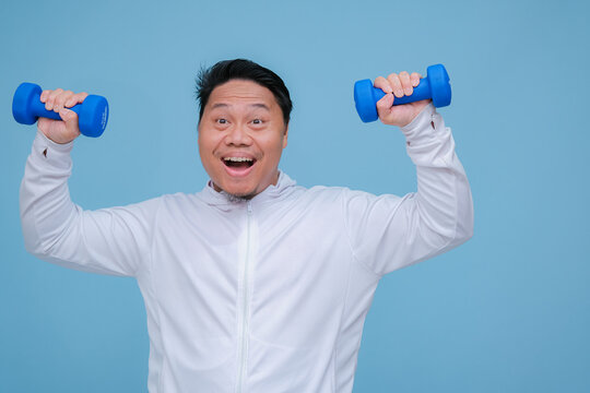 Close up of young Asian man in the gym lifting dumbbell wearing white t-shirt with happy laughing expression on turquoise blue background