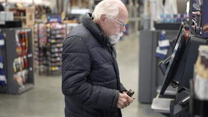 Senior man stands at self checkout in a hardware store completing his shopping. He holds his wallet while scanning items. High quality 4k footage
