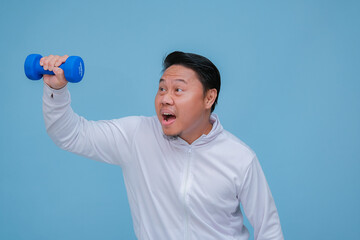 Close up of young Asian man in the gym lifting dumbbell wearing white t-shirt with happy laughing expression on turquoise blue background