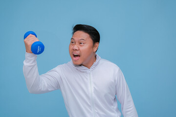 Close up of young Asian man in the gym lifting dumbbell wearing white t-shirt with happy laughing expression on turquoise blue background