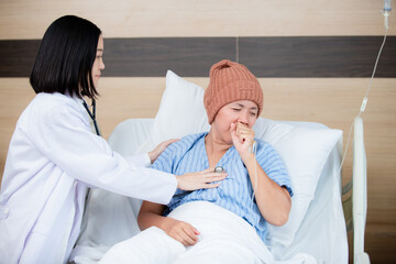 A compassionate doctor examines a female patient wearing a knitted hat in a hospital bed, checking her heartbeat with a stethoscope, showing care and medical attention during treatment.