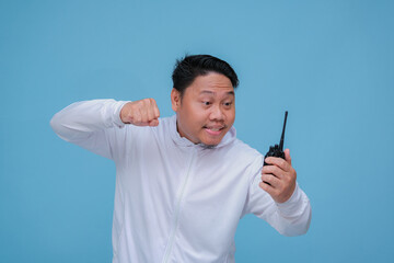 Young Asian man talking on Handy Talkie with very angry expression wearing white t-shirt on turquoise blue backgroun