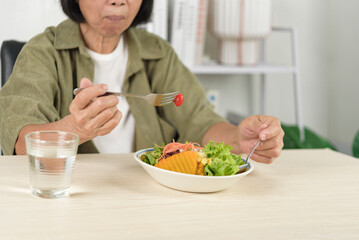 Senior Asian woman eating fresh vegetable salad with pumpkin and drinking water at home. Elderly healthy lifestyle, organic nutrition, weight loss, and vegetarian diet concept.
