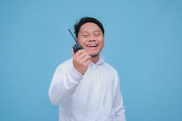 Closeup of Asian man talking on Handy Talky with laughing expression and pointing finger wearing white t-shirt on turquoise blue background 