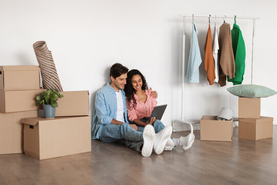 Cheerful couple sits on the floor with moving boxes around them, using a digital tablet to find furniture and household items for their new home. They look excited and engaged in their search. - Powered by Adobe