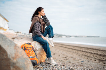 Young woman sitting on coastal rocks with orange backpack, resting by the sea and enjoying peaceful seaside atmosphere during relaxed outdoor travel moment
