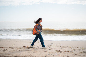 Woman walking along empty sandy beach with orange backpack, solo travel lifestyle portrait, calm coastal atmosphere, freedom, independence, mindfulness, ocean waves, minimal scenery