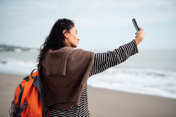 Woman with backpack taking selfie on sandy beach near ocean waves, modern travel lifestyle, solo tourism concept, digital communication, freedom, technology, seaside vacation