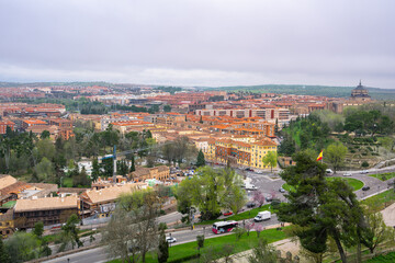 A high angle view of the historic architecture and orange rooftops of Toledo, Spain, with lush green trees under a soft overcast sky.
