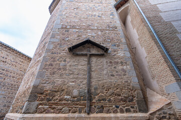 A large wooden cross mounted on the historic stone masonry walls of Toledo city under a cloudy spring sky in Spain.