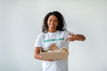 An African American female volunteer holds a box filled with food donations. She points at the box...