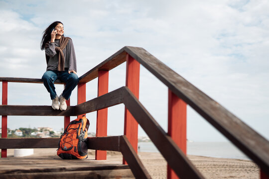 Young adult woman sitting on wooden beach stairs, talking on smartphone, casual striped clothing, backpack nearby, relaxed mood, seaside environment, cloudy sky, natural daylight.