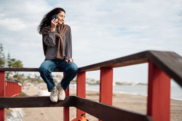 Young adult woman sitting on wooden beach stairs, talking on smartphone, casual striped clothing, backpack nearby, relaxed mood, seaside environment, cloudy sky, natural daylight.