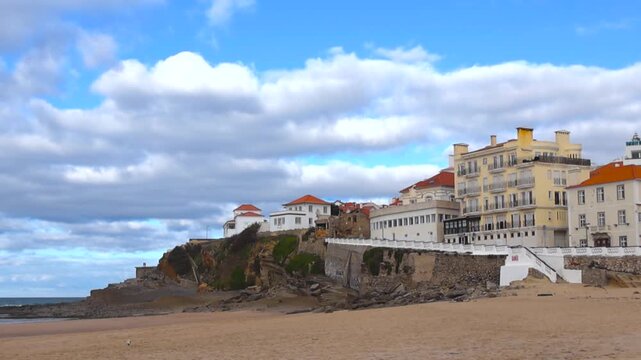 Pan shot of Praia das Ma&ccedil;&atilde;s coastline in Portugal, showing white cliffside houses, sandy beach and Atlantic waves crashing on rocky shore under blue sky with clouds, no people visible