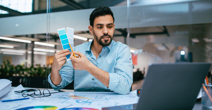 A man is engaged in selecting color shades from a palette while sitting at a desk. He is looking at a laptop and has design materials spread out in front of him in a bright office space. - Powered by Adobe