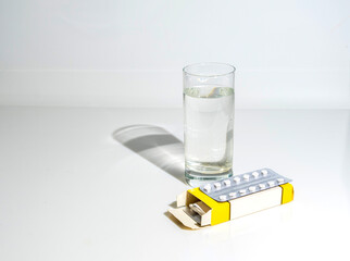 A box of pills and a glass of water on white glass, with shadow, copy space