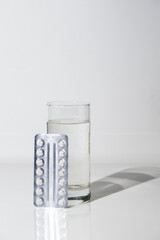 A blister pack of white tablets rests on a glass of water, on white glass, space for text