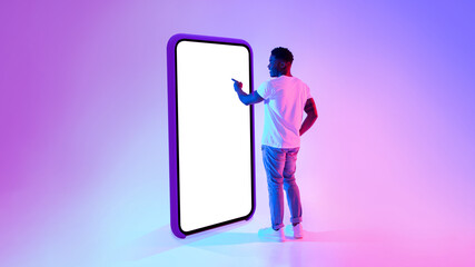 A black man stands next to a large smartphone mockup, engaged in exploring the user interface under vibrant neon lights. This scene showcases digital interaction in a modern setting.