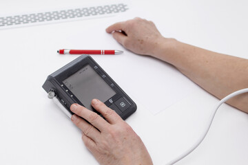 Close-up of elderly woman using digital blood pressure monitor and writing health data on white paper at home, concept of self-monitoring and hypertension control.