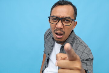 Portrait of Asian man pointing with his index finger forward, angry expression on a blue background.