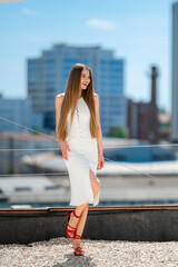 Caucasian Woman In White Buttoned Midi Dress Posing On Rooftop With City Skyline Background, Red Strappy