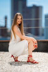 Caucasian Woman Crouching On Rooftop Gravel Adjusting Red Heels While Wearing White Dress, Poised Fashion Pose
