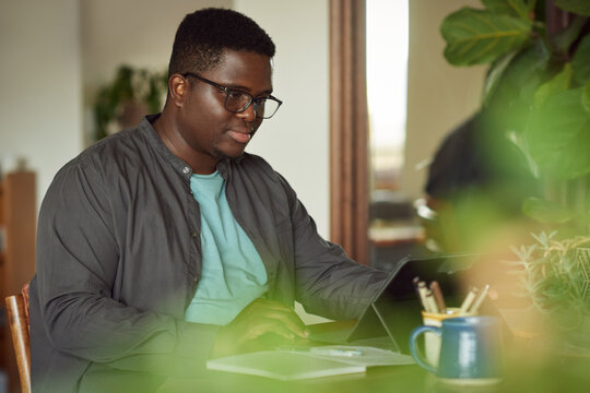 Young African man sitting at home and working remotely using a tablet