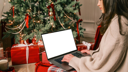 Woman using laptop near decorated Christmas tree surrounded by gifts and candles