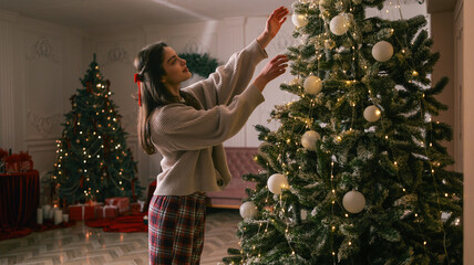Young woman decorating a Christmas tree with ornaments and lights in a cozy room
