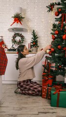 Woman decorating a Christmas tree with red ornaments in a cozy kitchen setting