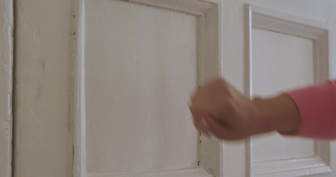 Close up of a hand wearing a pink sleeve knocking on a white wooden door, conveying an element of interaction. Slow motion.