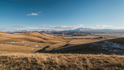 Open plain landscape with rolling hills, mountain range, and snow patches under a blue sky.