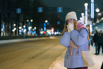 Young woman standing on a snowy sidewalk at a bus stop during a cold winter night, dressed in warm clothing and blowing on her hands for warmth while waiting for public transportation in the city