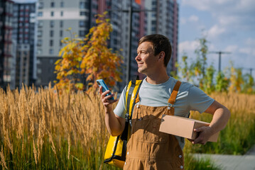 Young male courier delivering package in urban residential area, checking delivery app on smartphone, providing fast and reliable service to customers outdoors in city