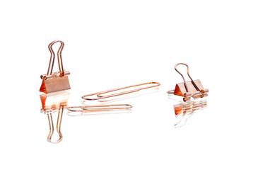 Copper binder clips and paper clips arranged on a white reflective surface. Minimal office supplies concept with clean composition and metallic texture.