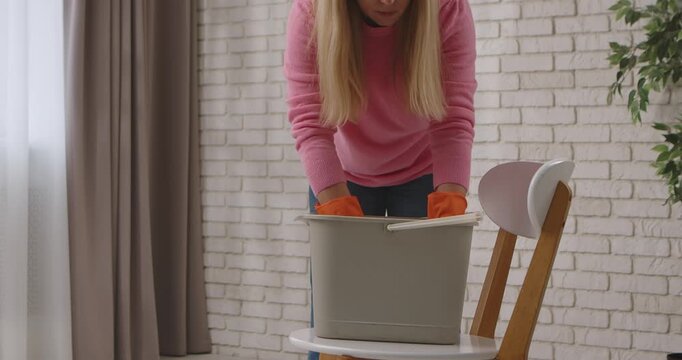 A woman in a pink sweater and orange gloves rinses a mop cloth in a bucket of water in a well lit room. Slow motion.