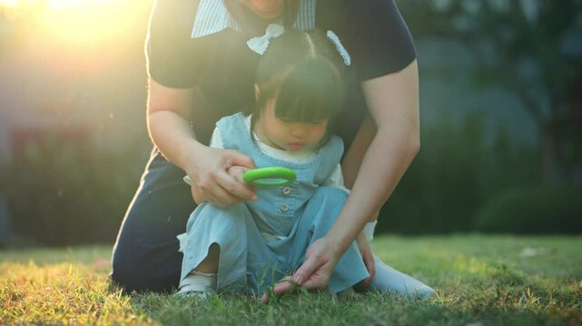 Happy Asian mother and her little toddler daughter exploring nature together with a magnifying glass on the grass in a sunny outdoor park