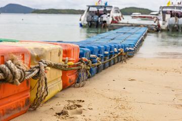 Colorful floating plastic dock form a long tethered line along a sandy beach, leading to boats at a calm harbor. Bright hues and ropes create a vivid nautical scene with a summer vibe. © Anna