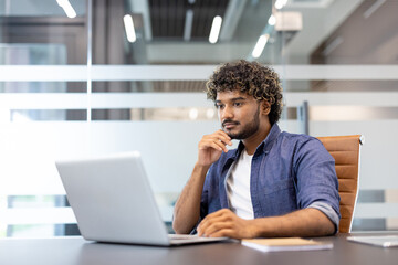 Serious young thoughtful Indian man working in the office on a laptop