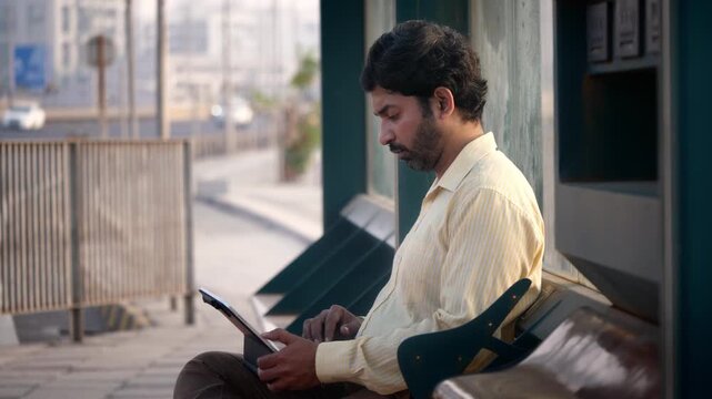 A daylight outdoor shot of a middle aged Indian Asian corporate office man or male freelancer waiting at a bus stop, busy using and typing on a laptop to complete the office assignment.