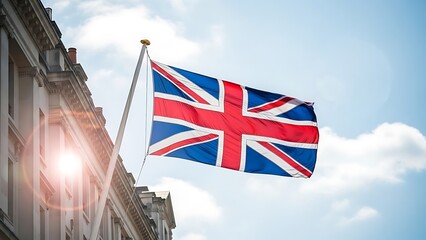 Union jack flag of the united kingdom waving on a flagpole against a bright blue sky with lens flare from a nearby building facade