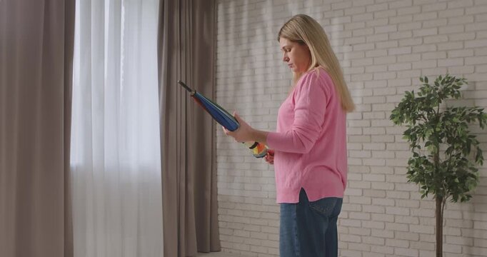 A woman in a pink sweater begins to open a folded rainbow umbrella while standing in a bright room with soft lighting. Slow motion.