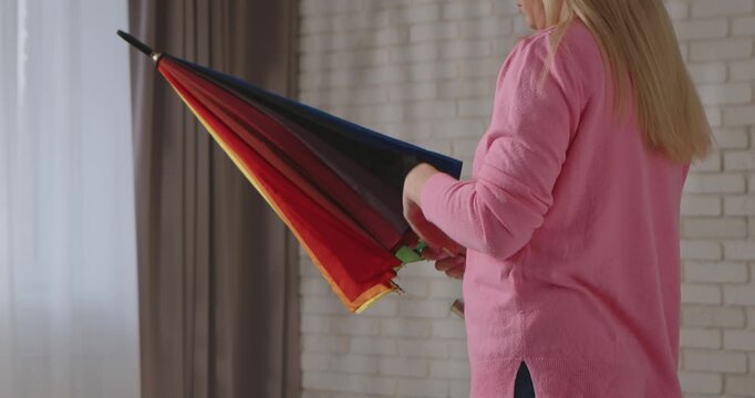 A woman in a pink sweater carefully folds a colorful rainbow umbrella while standing in a bright room with soft natural lighting. Slow motion.