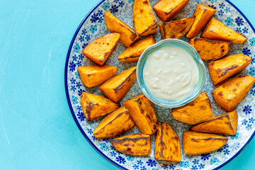 Top view of a plate with roasted sweet potatoes. Small bowl with lemon-tahini sauce in the center. Plain blue background.